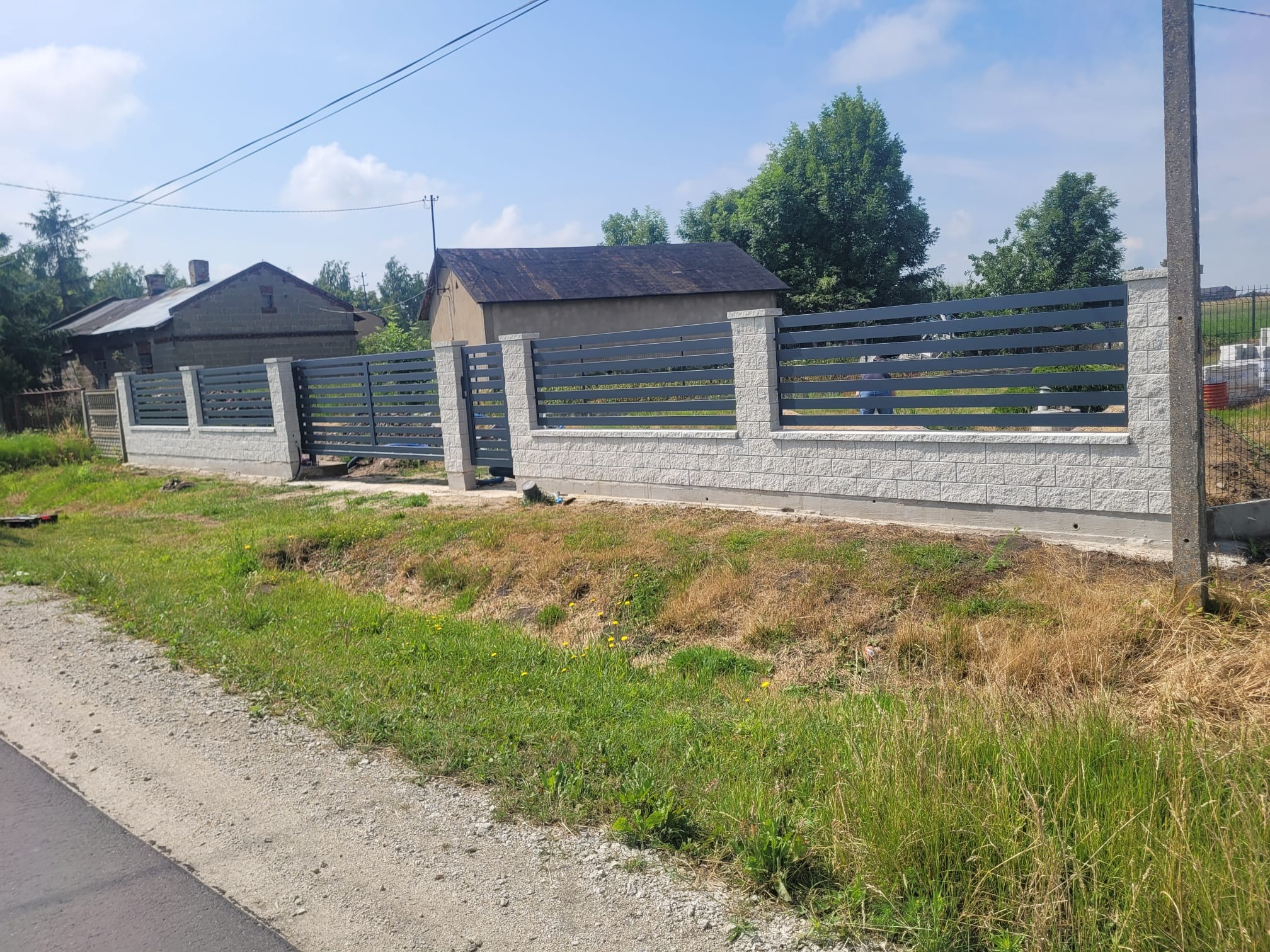 Nowoczesne ogrodzenie posesji z szarymi, poziomymi panelami i białymi, murowanymi słupkami. Widok na dom w tle, trawa i droga na pierwszym planie.