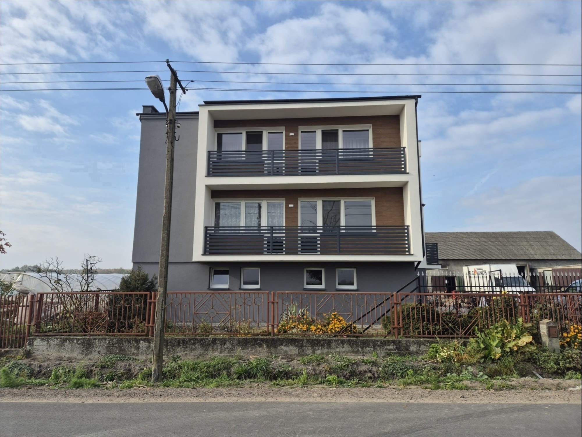 Dwukondygnacyjny dom z balkonami z poziomymi, szarymi barierkami. W tle błękitne niebo z chmurami. Na pierwszym planie metalowe ogrodzenie posesji.