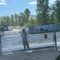 Tortura Rdzy piaskowanie, malowanie proszkowe, konserwacje - Pracownik w kombinezonie ochronnym i hełmie piaskuje metalową bramę wjazdową na posesję, widoczne pylenie i otwarte skrzydło bramy, w tle budynki i drzewa.