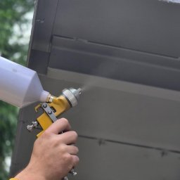 Tortura Rdzy piaskowanie, malowanie proszkowe, konserwacje - Szary metalowy element konstrukcyjny natryskiwany farbą z żółtego pistoletu lakierniczego, widoczna ręka w żółtej rękawicy ochronnej i przewód doprowadzający farbę.