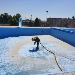 Tortura Rdzy piaskowanie, malowanie proszkowe, konserwacje - Pracownik w kombinezonie i kasku piaskuje dno pustego, błękitnego basenu na zewnątrz, z widoczną zjeżdżalnią i budynkami w tle.