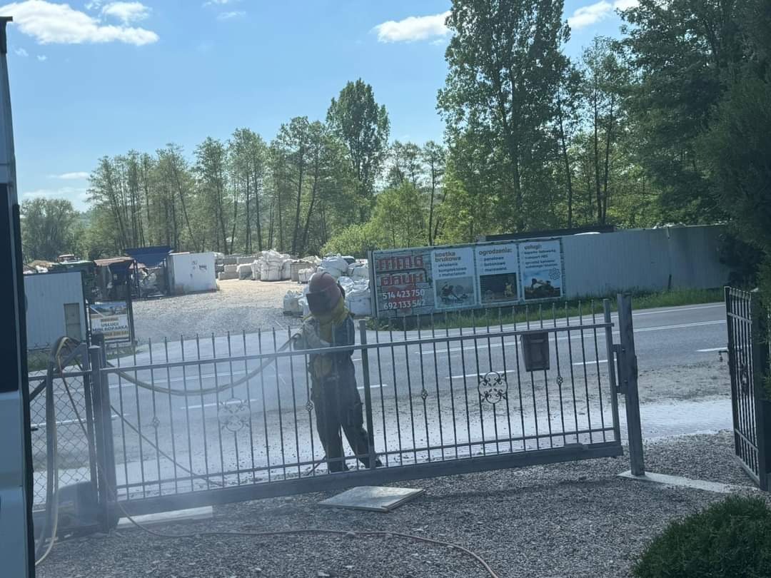 Pracownik w kombinezonie ochronnym i hełmie piaskuje metalową bramę wjazdową na posesję, widoczne pylenie i otwarte skrzydło bramy, w tle budynki i drzewa.