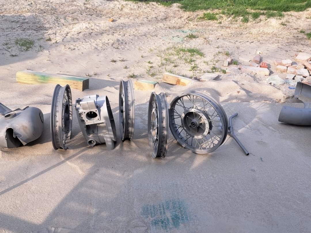Elementy motocykla, w tym koła, silnik i zbiornik paliwa, przygotowane do malowania proszkowego, rozłożone na piaszczystym podłożu w plenerze.