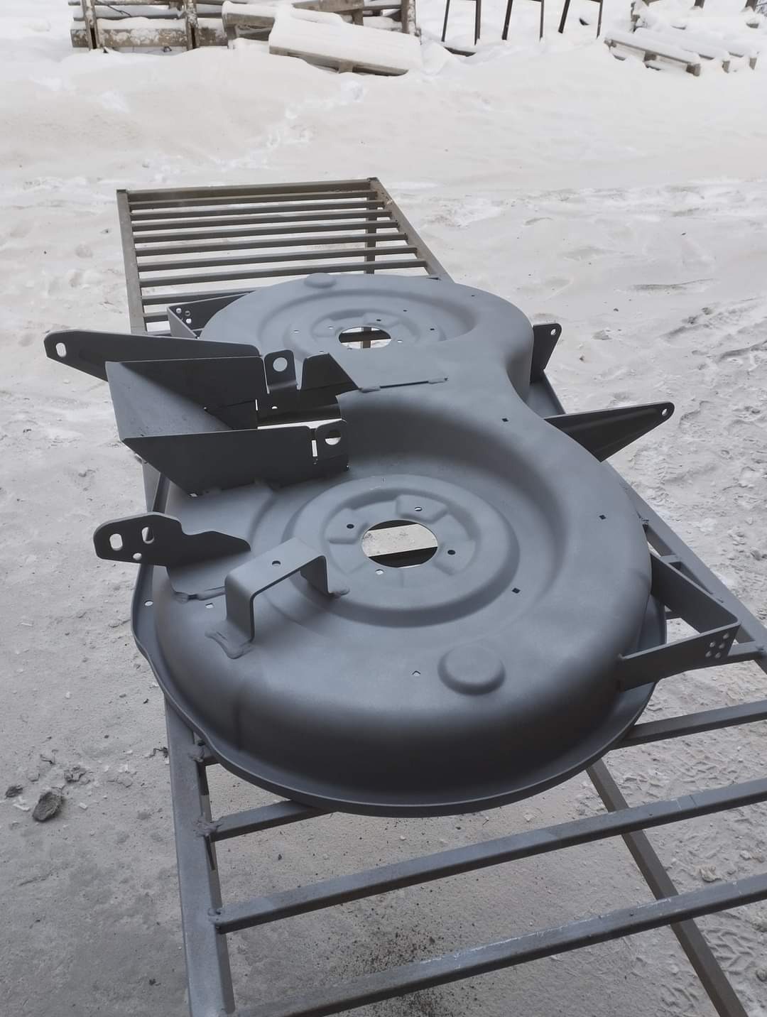 Szary element obudowy kosiarki spalinowej po piaskowaniu i malowaniu na metalowym ruszcie, w tle śnieg.