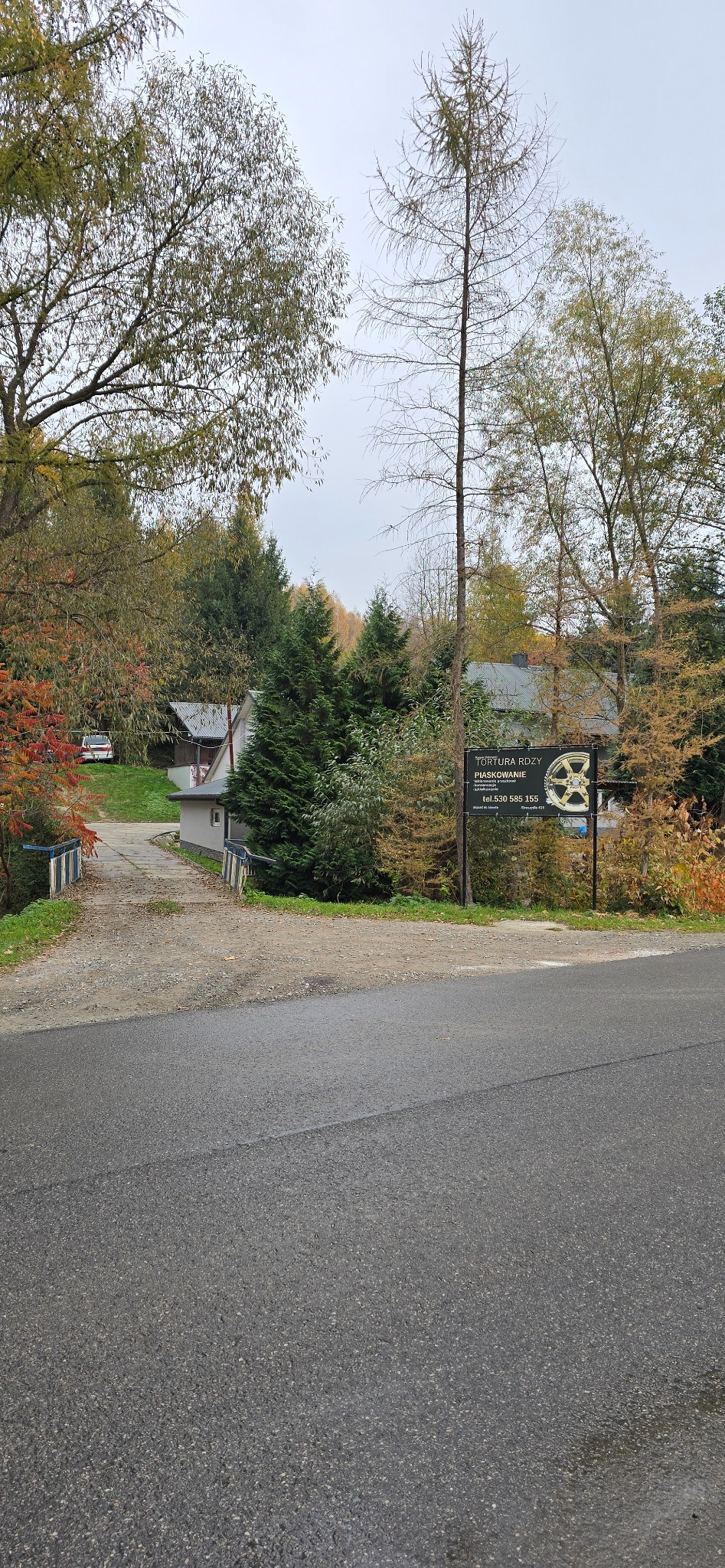 Tablica reklamowa firmy oferującej usługi piaskowania, z logo przedstawiającym felgę, na tle jesiennego krajobrazu z drzewami i budynkami.