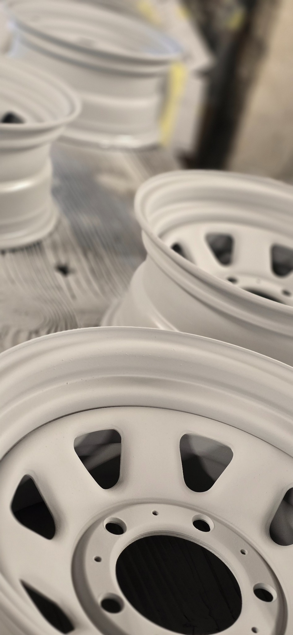 Pomalowane na biało, odnowione felgi samochodowe z widoczną fakturą proszkową, ułożone na drewnianej powierzchni, z rozmytym tłem warsztatowym.
