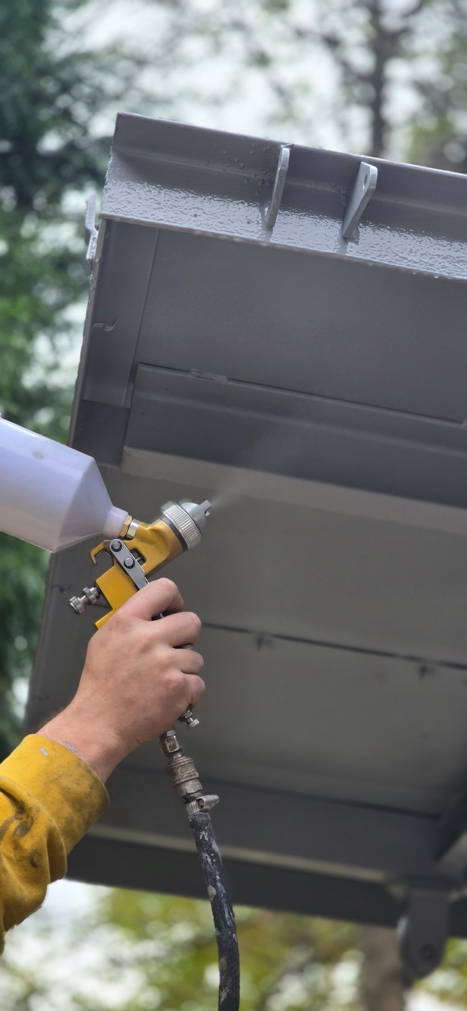 Szary metalowy element konstrukcyjny natryskiwany farbą z żółtego pistoletu lakierniczego, widoczna ręka w żółtej rękawicy ochronnej i przewód doprowadzający farbę.
