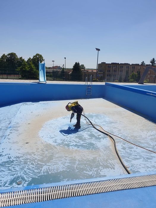 Pracownik w kombinezonie i kasku piaskuje dno pustego, błękitnego basenu na zewnątrz, z widoczną zjeżdżalnią i budynkami w tle.