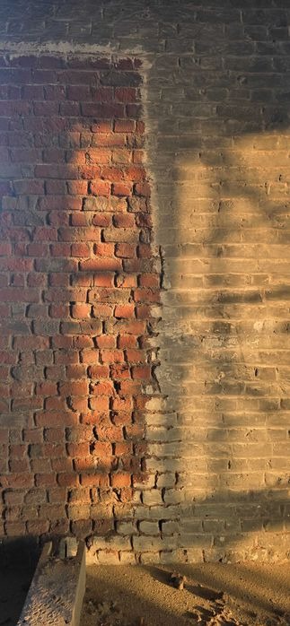 Fragment ceglanej ściany po czyszczeniu, z widocznym podziałem na obszar oczyszczony i nieoczyszczony, różniące się kolorem i strukturą, oświetlony ciepłym światłem.