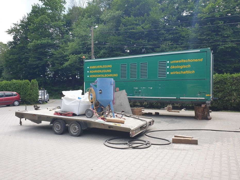 Przyczepa z piaskarką ciśnieniową, workiem ścierniwa i wężami na tle zielonego kontenera z hasłami ekologicznymi i informacjami o usługach.