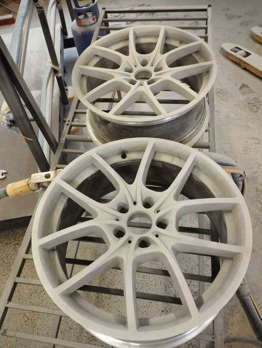 Dwa aluminiowe felgi samochodowe, przygotowane do malowania proszkowego, ustawione jeden na drugim na metalowym regale w warsztacie.