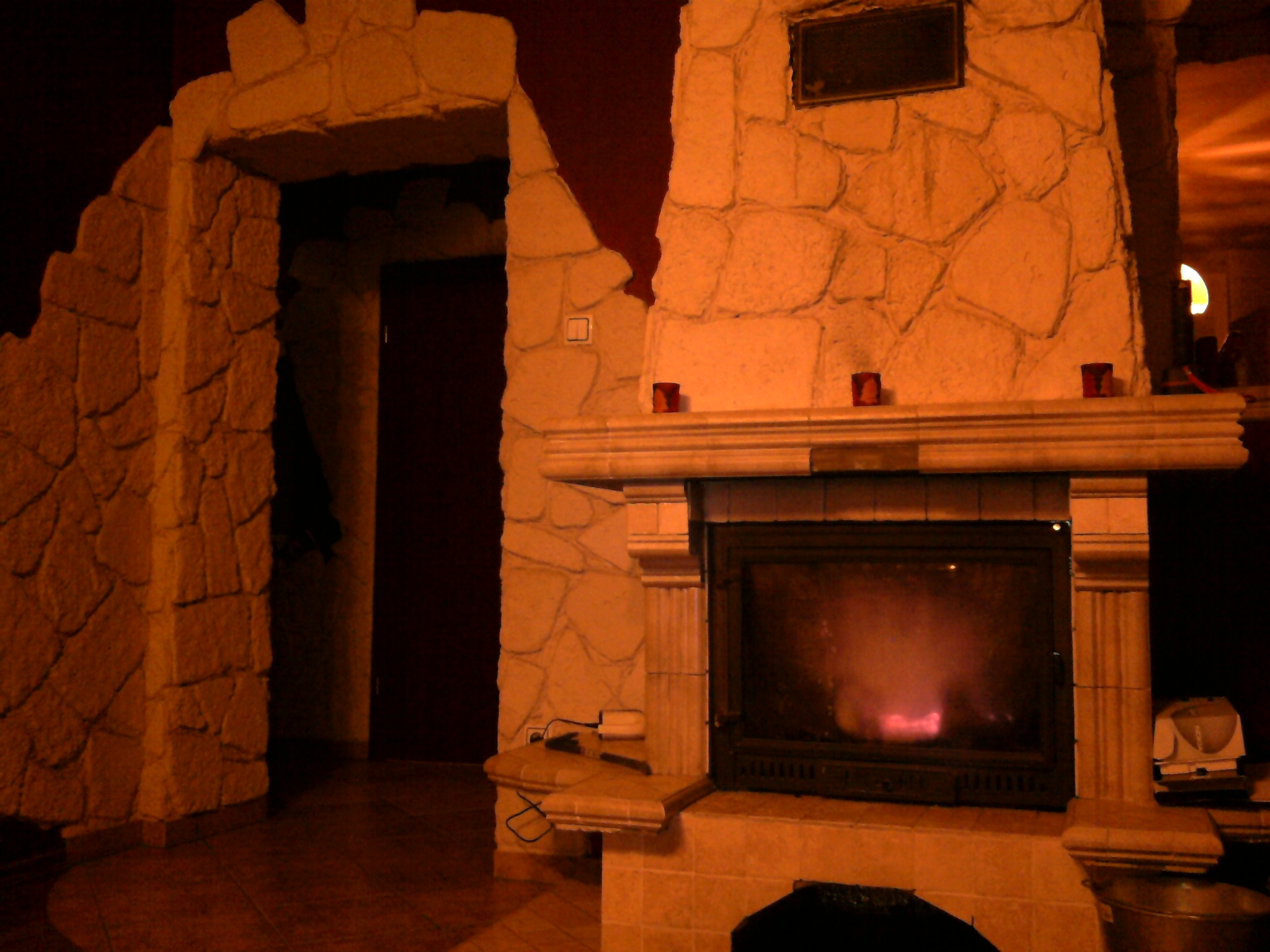 Kamienny portal wejściowy i kominek z paleniskiem, oświetlone ciepłym światłem, z elementami dekoracyjnymi jak świeczniki i kamienna obudowa, tworzące przytulny wystrój wnętrza.