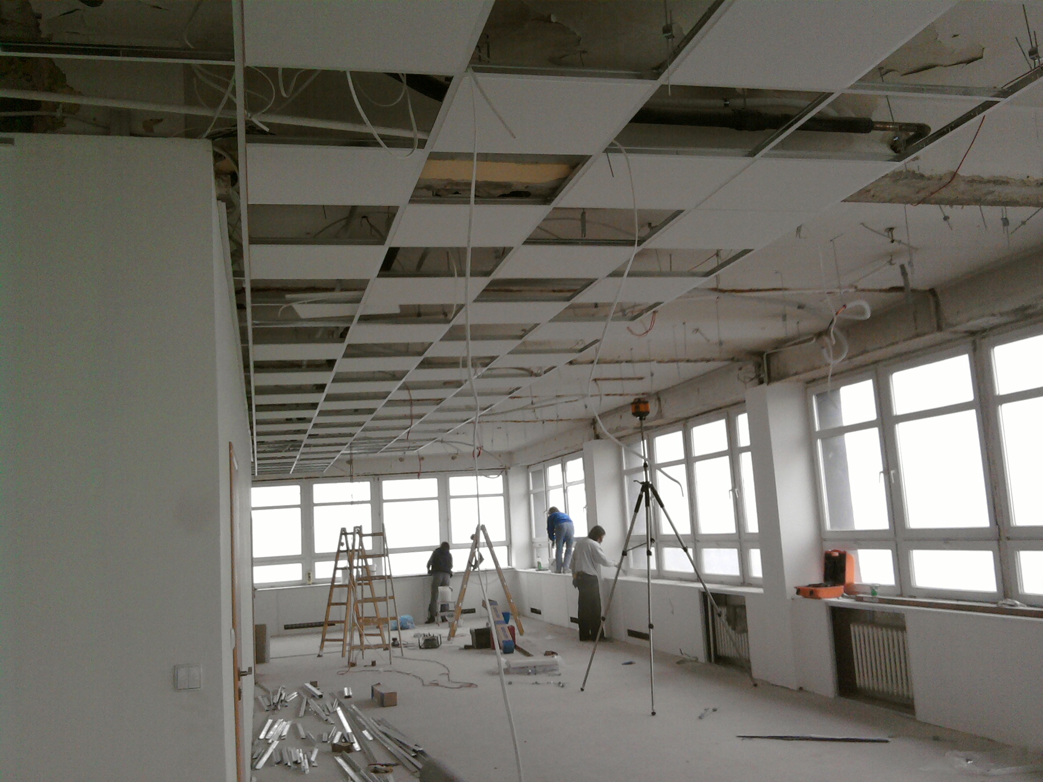 Montaż sufitu podwieszanego w dużym, jasnym pomieszczeniu z oknami; widoczne drabiny, profile i poziomica laserowa na statywie.