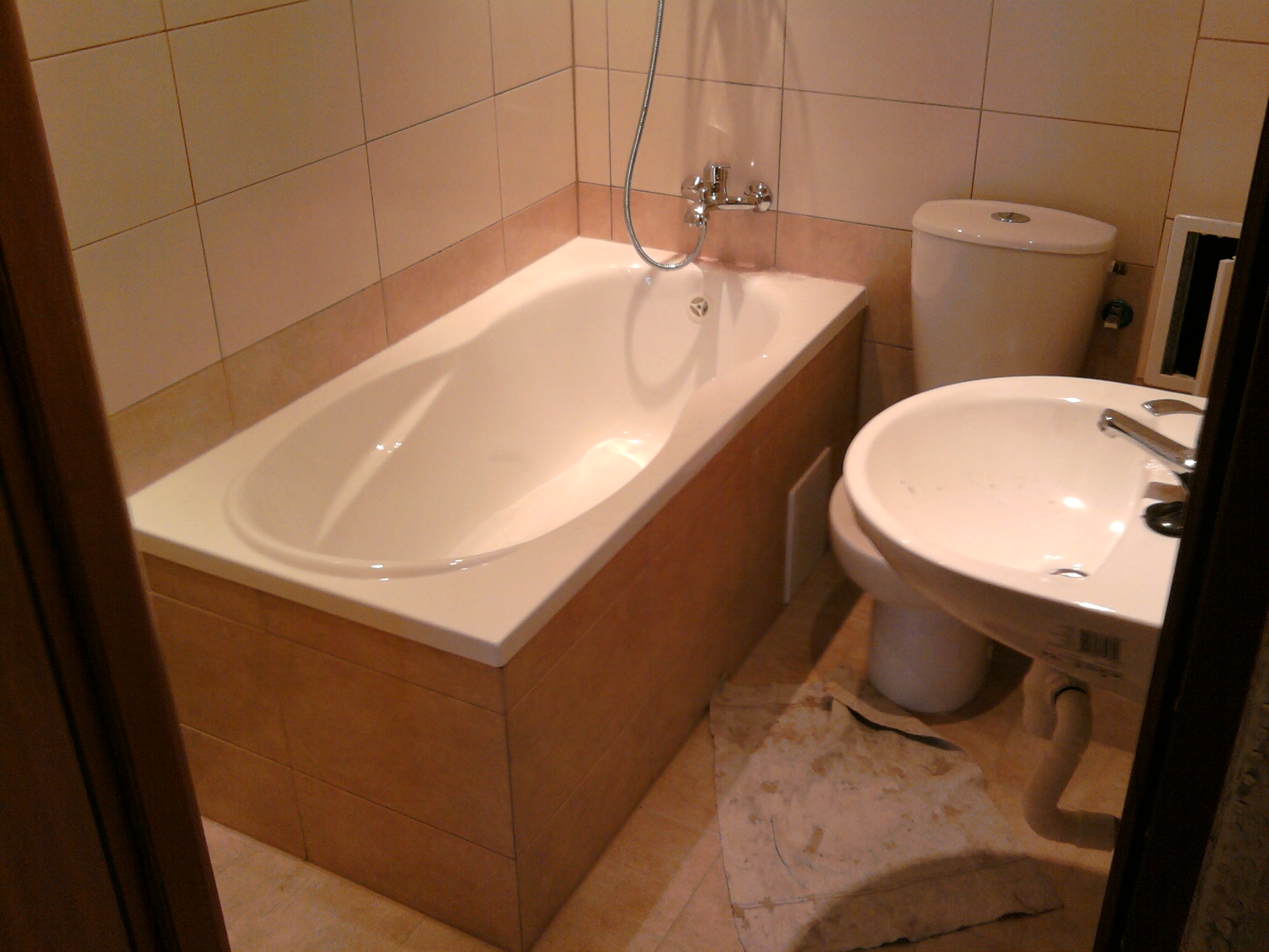 Wykończona łazienka z beżowymi płytkami, obejmująca wannę z obudową z płytek, umywalkę na postumencie i toaletę. Na podłodze widoczne resztki po remoncie.