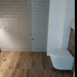 Remont łazienki w trakcie: ułożone płytki ścienne w odcieniach szarości z widocznymi otworami na armaturę, drewnopodobna podłoga i wisząca toaleta w nowoczesnym stylu.