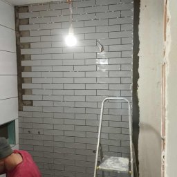 Kacper bud - Układanie szarych, podłużnych płytek w jodełkę na ścianie łazienki, widoczna drabina aluminiowa i wisząca żarówka oświetlająca fragment ściany z płytkami, plastikowe krzyżyki dystansowe między...