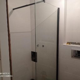 Kacper bud - Szklana kabina prysznicowa z czarnymi profilami w łazience wyłożonej białymi płytkami, widoczny panel sterujący spłuczką i poziomica na półce.