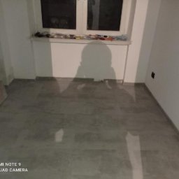 Kacper bud - Pusty pokój z szarą podłogą i białymi ścianami, widoczne narzędzia na parapecie okna, cień osoby robiącej zdjęcie.