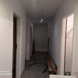 Kacper bud - Korytarz w trakcie malowania na biało, widoczne świeżo pomalowane ściany, surowa podłoga, listwy przypodłogowe w kartonie i przewody na podłodze, oświetlenie sufitowe punktowe.