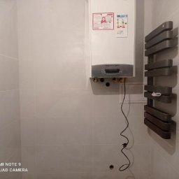 Kacper bud - Biały piec gazowy z szarym panelem i etykietami informacyjnymi zamontowany na ścianie z jasnych płytek, obok grzejnik drabinkowy w odcieniach szarości, widoczne przewody instalacji hydraulicznej...