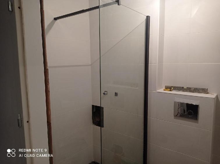 Zamontowana kabina prysznicowa z czarnymi profilami w łazience wyłożonej białymi, prostokątnymi płytkami; widoczny poziomiczka i narzędzia na półce.