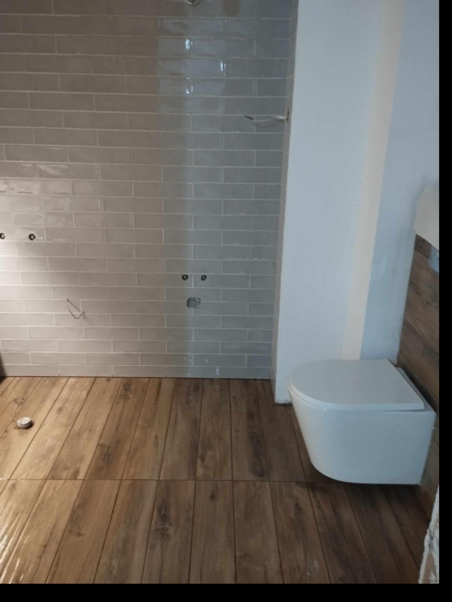 Remont łazienki w trakcie: ułożone płytki ścienne w odcieniach szarości z widocznymi otworami na armaturę, drewnopodobna podłoga i wisząca toaleta w nowoczesnym stylu.