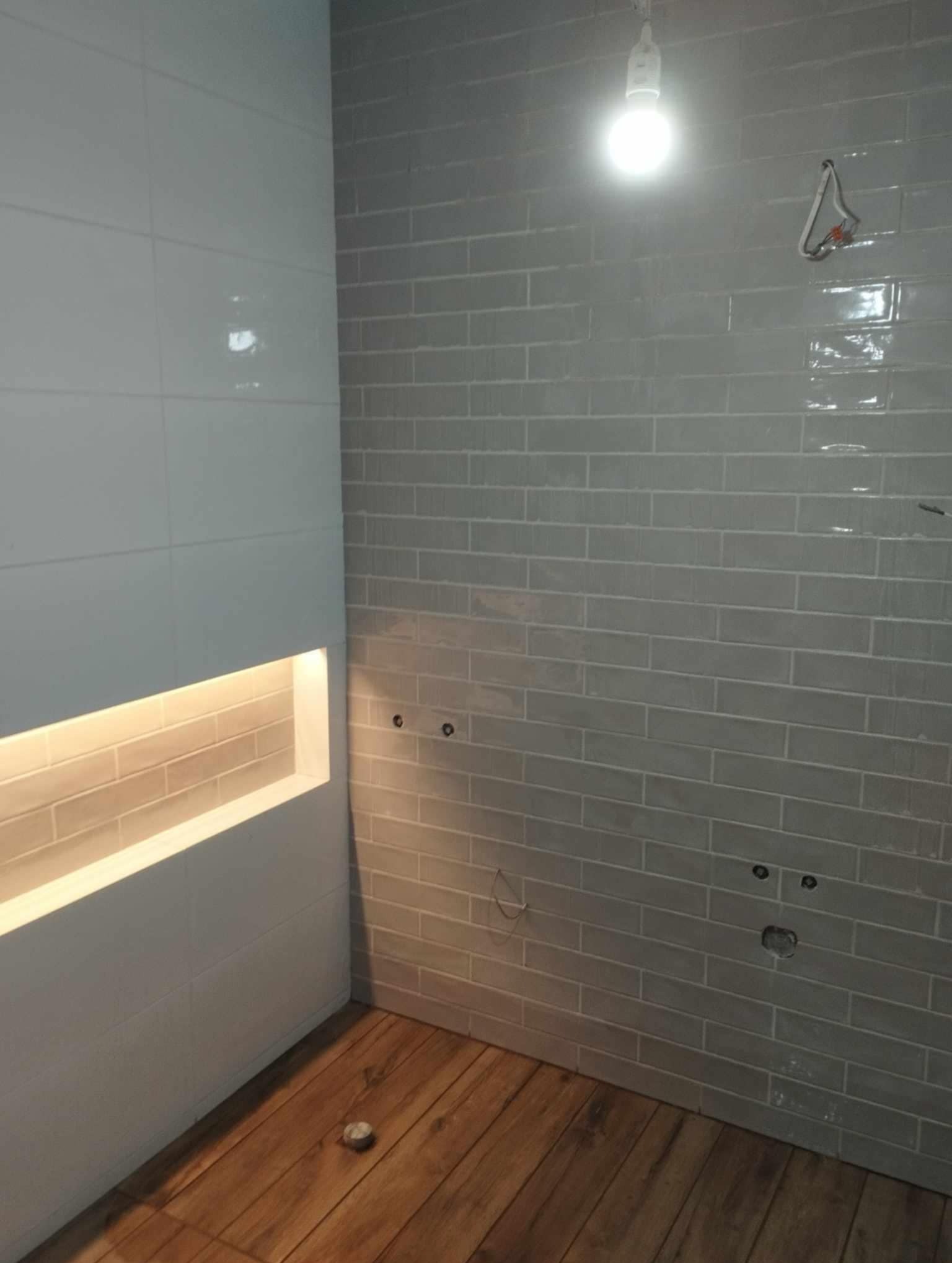 Świeżo wykafelkowana ściana w łazience z szarymi, podłużnymi płytkami, drewnopodobna podłoga, wnęka z podświetleniem i wystające kable.
