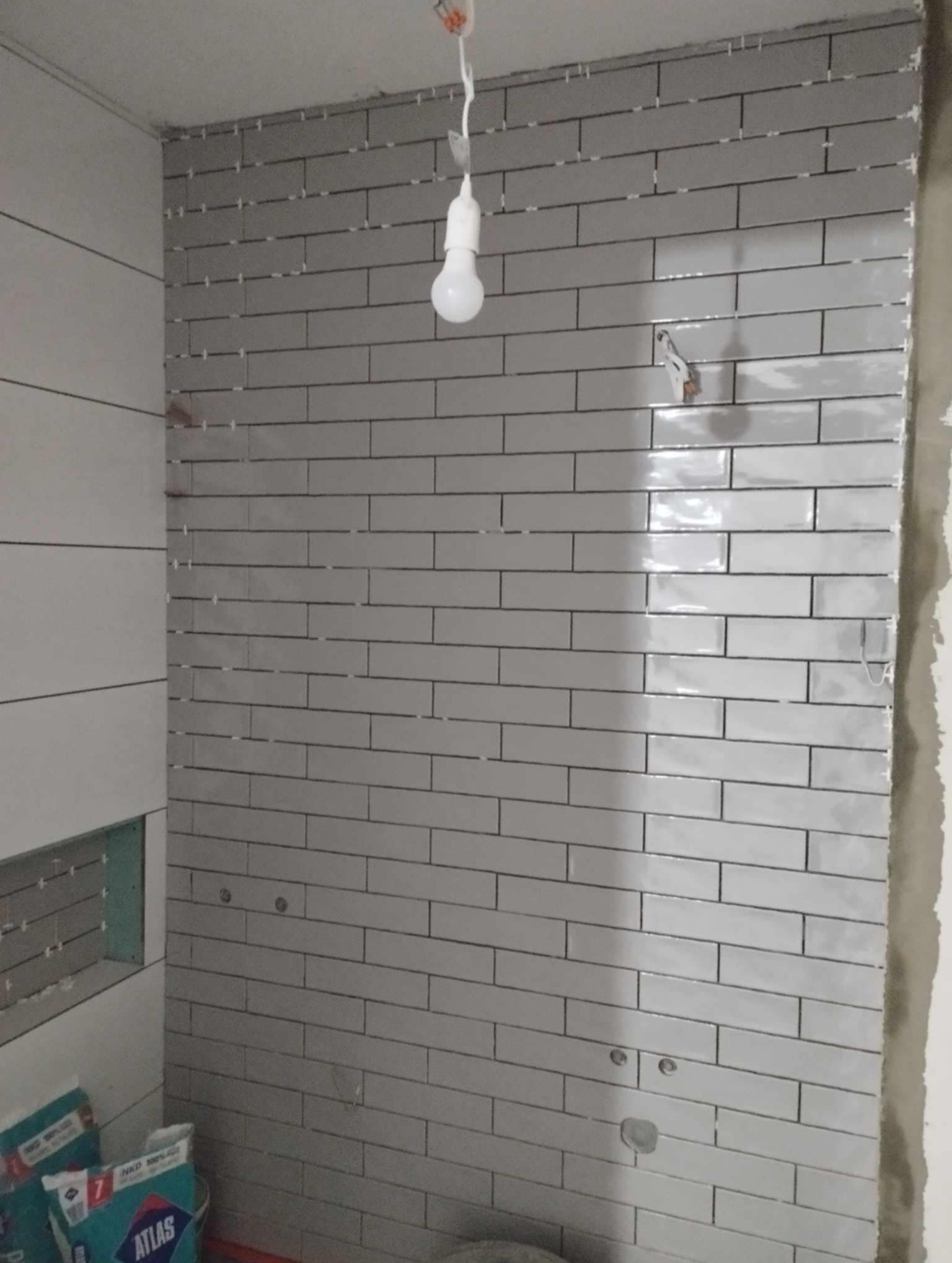 Ściana łazienki w trakcie układania szarych, podłużnych płytek, widoczne krzyżyki dystansowe, surowy tynk, wisząca żarówka i worki z klejem do płytek w dolnym rogu.