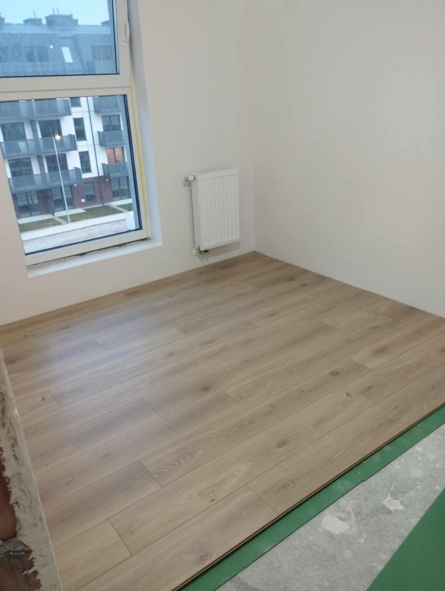 Fragment pokoju z nową, jasną podłogą laminowaną, częściowo ułożoną na zielonym podkładzie, widok na blok mieszkalny za oknem.