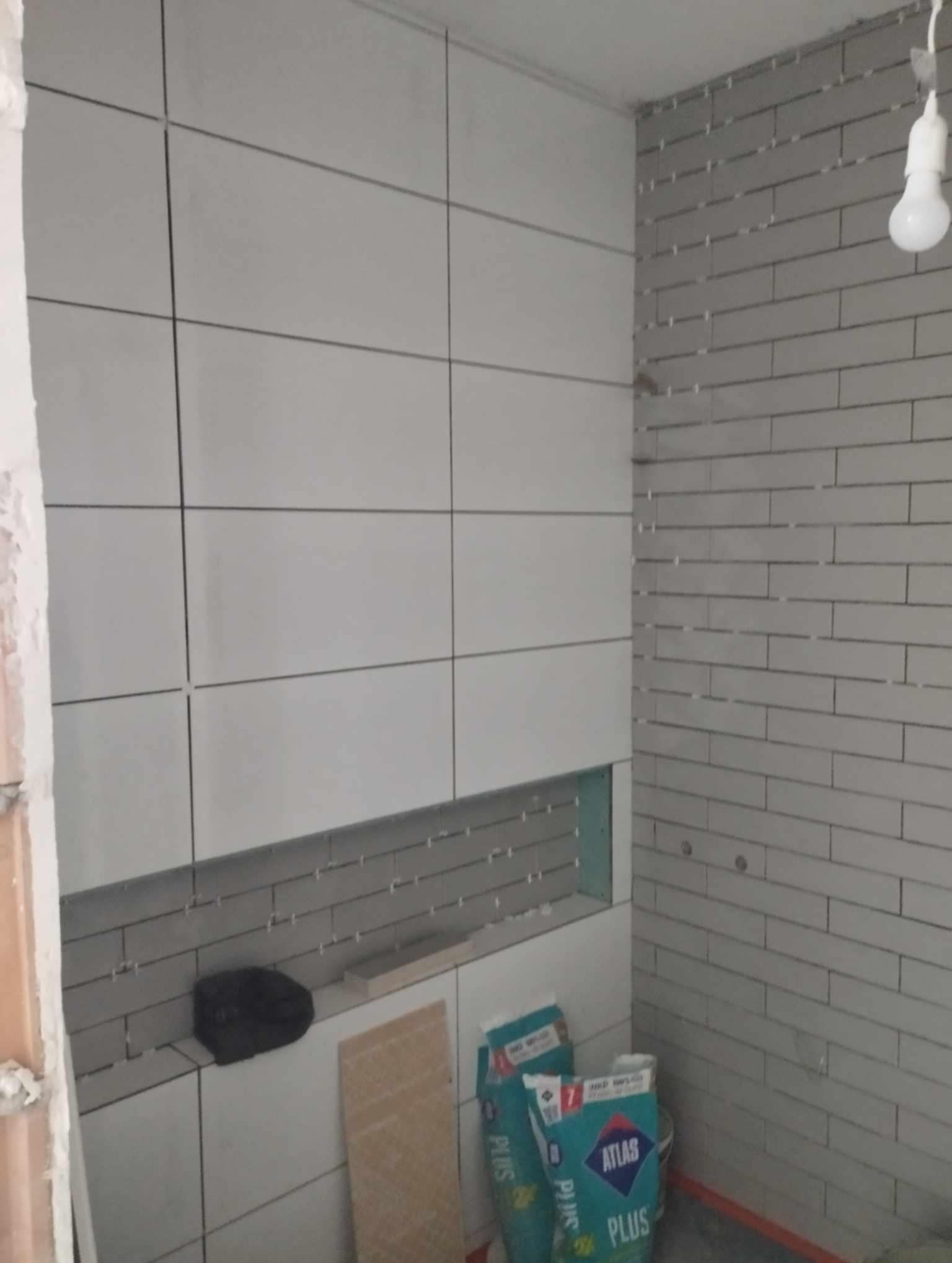 Ściana łazienki w trakcie układania płytek: duże, jasne płytki na jednej ścianie i małe, szare płytki na drugiej, widoczne worki z klejem Atlas Plus, niezabudowana wnęka i wisząca żarówka.