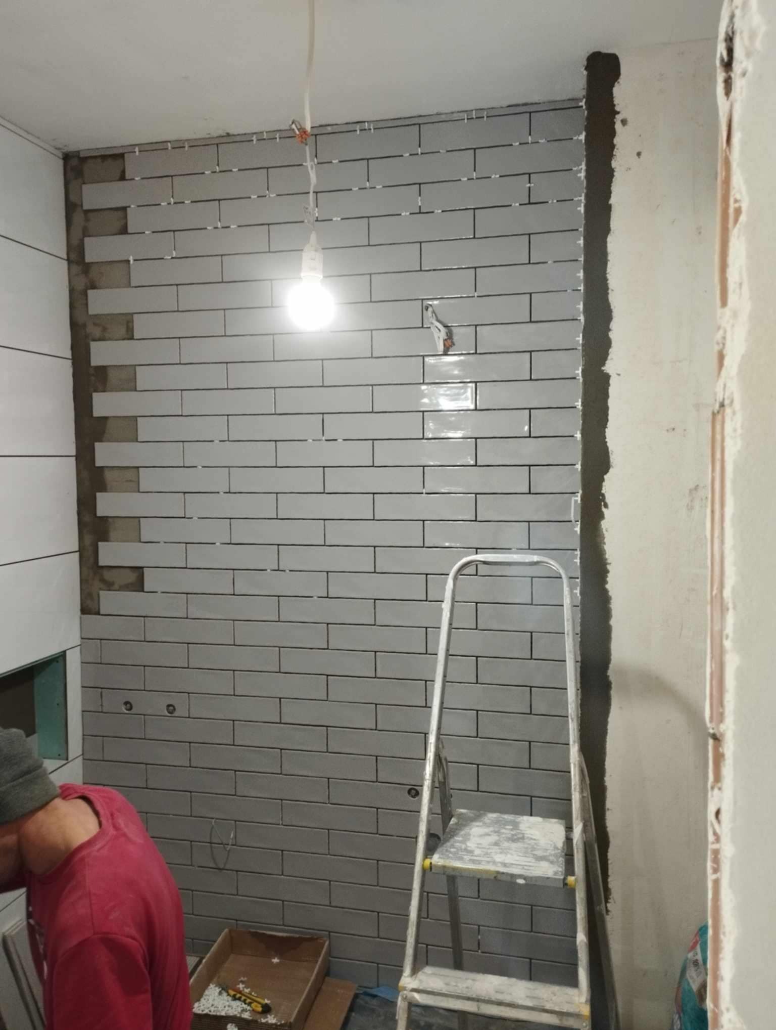 Układanie szarych, podłużnych płytek w jodełkę na ścianie łazienki, widoczna drabina aluminiowa i wisząca żarówka oświetlająca fragment ściany z płytkami, plastikowe krzyżyki dystansowe między...