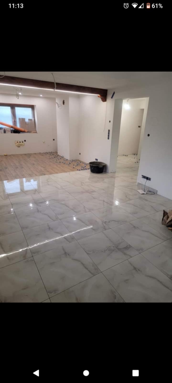 Układanie płytek podłogowych imitujących marmur w jasnym, przestronnym wnętrzu z drewnianym sufitem i widocznymi elementami instalacji elektrycznej oraz fragmentem podłogi bez płytek z rozsypanymi...