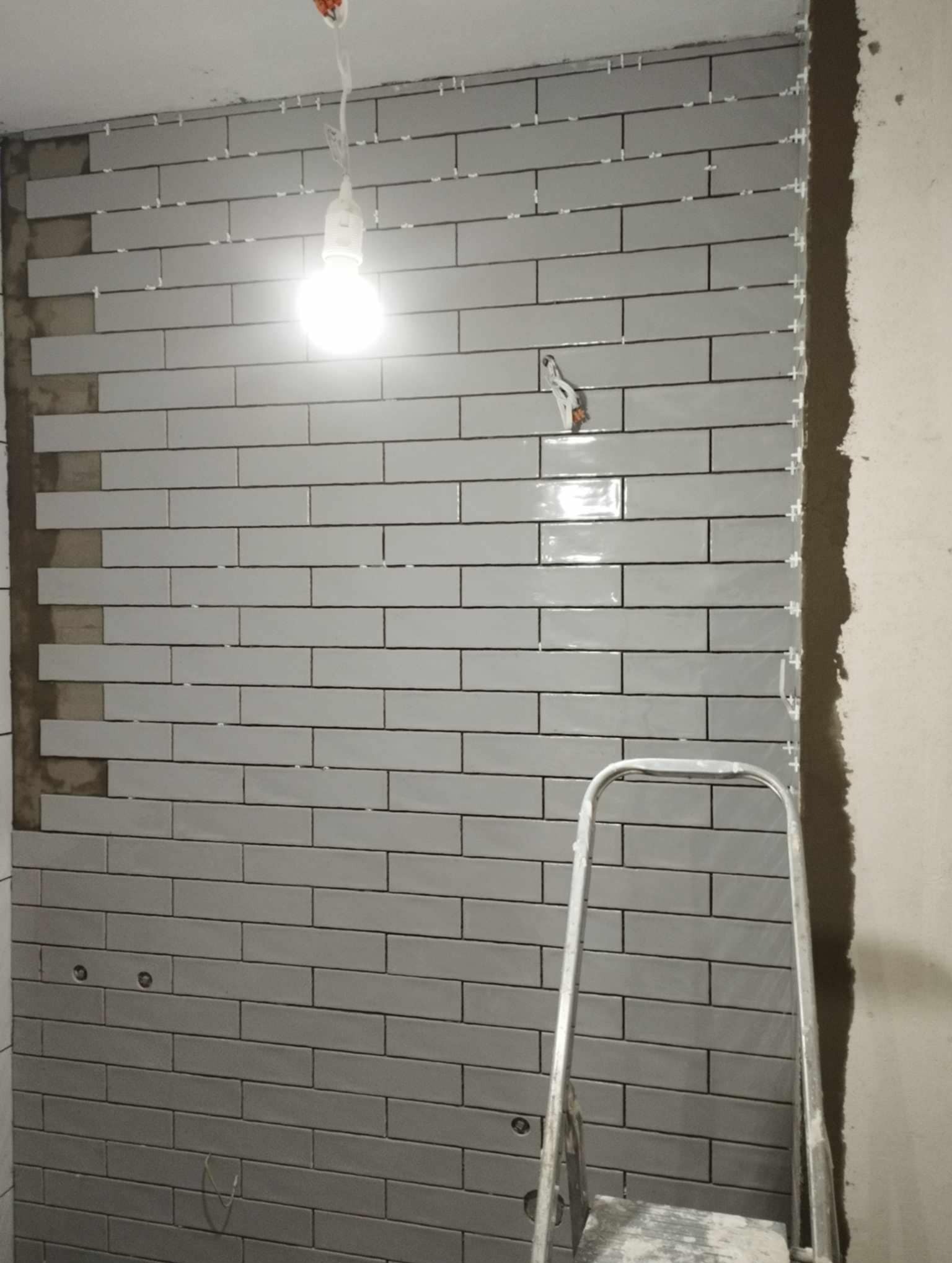 Ściana łazienki w trakcie układania szarych, podłużnych płytek z czarną fugą, widoczne krzyżyki dystansowe i drabina.