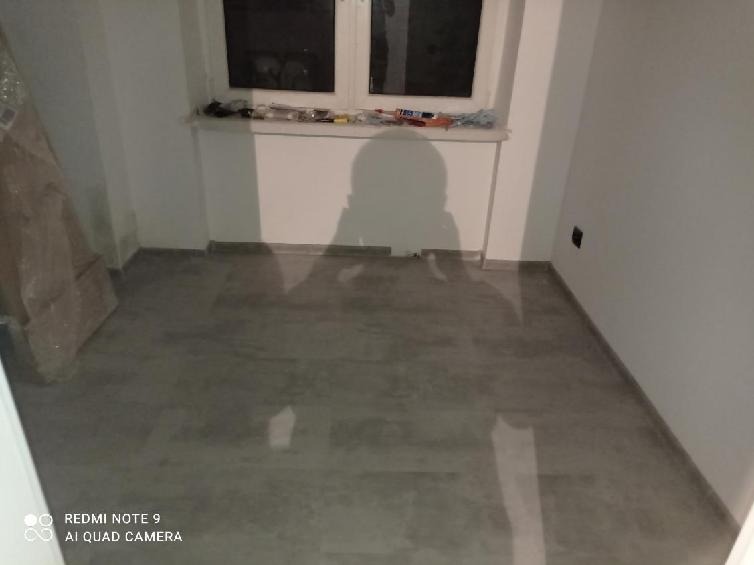 Pusty pokój z szarą podłogą i białymi ścianami, widoczne narzędzia na parapecie okna, cień osoby robiącej zdjęcie.