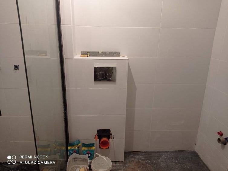 Montaż stelaża podtynkowego WC w łazience, widoczne białe płytki, poziomica na stelażu, przyłącze kanalizacyjne i worki z zaprawą klejową.