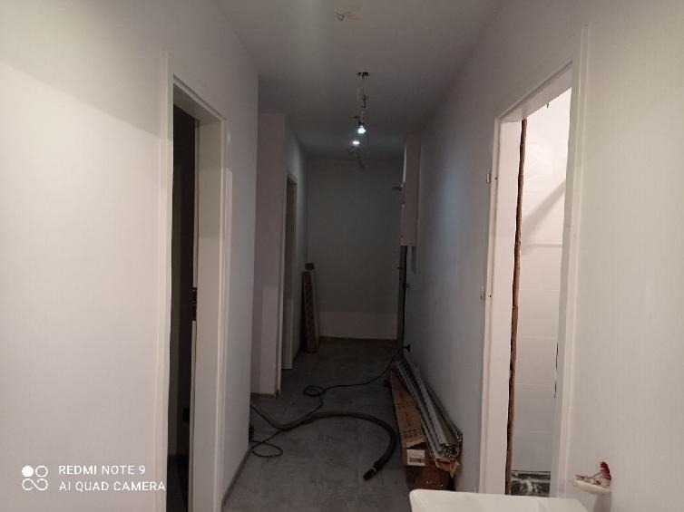 Korytarz w trakcie malowania na biało, widoczne świeżo pomalowane ściany, surowa podłoga, listwy przypodłogowe w kartonie i przewody na podłodze, oświetlenie sufitowe punktowe.