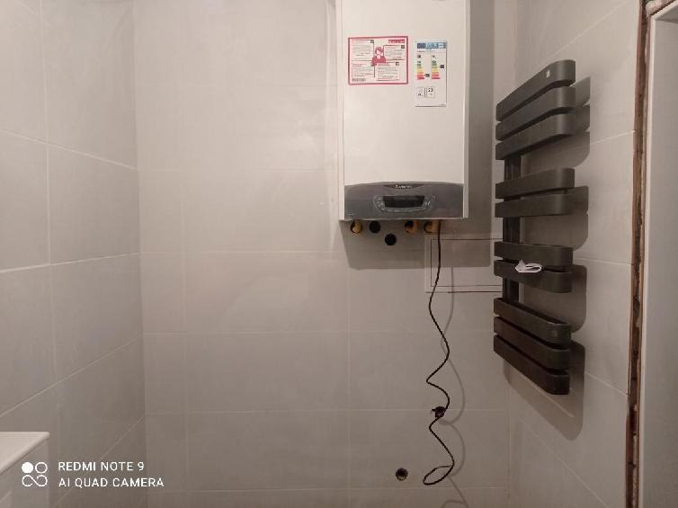 Biały piec gazowy z szarym panelem i etykietami informacyjnymi zamontowany na ścianie z jasnych płytek, obok grzejnik drabinkowy w odcieniach szarości, widoczne przewody instalacji hydraulicznej...