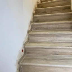 Schody wyłożone płytkami imitującymi drewno, widoczne ślady prac na ścianie, w tle fragment starej tapety.