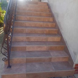 ART-HOUSE - Schody zewnętrzne wyłożone brązowymi płytkami, z ozdobną metalową balustradą, widoczne ślady prac wykończeniowych u podstawy.