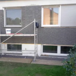 Montaż elewacji z szarej cegły klinkierowej na ścianie domu jednorodzinnego, widoczne rusztowanie, okna z roletami zewnętrznymi i fragment ogrodu z trawnikiem.