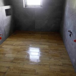 ART-HOUSE - Wykończona łazienka z szarymi płytkami ściennymi i drewnopodobną podłogą z płytek, małe okno w górnej części ściany, widoczne elementy instalacji wodnej.