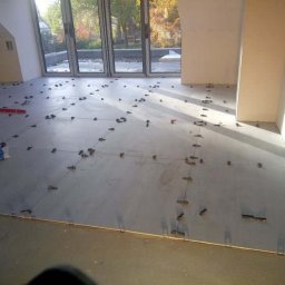 ART-HOUSE - Szare płytki podłogowe ułożone z użyciem klipsów poziomujących w jasnym pomieszczeniu z dużymi oknami balkonowymi, widoczne narzędzia glazurnicze.