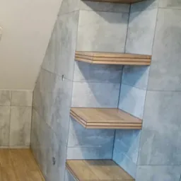 Wykończona łazienka na poddaszu z szarymi płytkami imitującymi beton i drewnianymi półkami wbudowanymi we wnękę