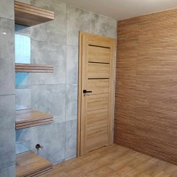 ART-HOUSE - Wykończone wnętrze: szare płytki imitujące beton na ścianie z wbudowanymi drewnianymi półkami, drewniane panele ścienne w odcieniach brązu oraz jasne drewniane drzwi z poziomymi frezowaniami.