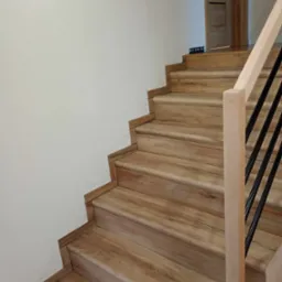 Schody drewniane z jasną balustradą i czarnymi prętami, wykończone listwami przypodłogowymi w kolorze drewna, widok z dołu.