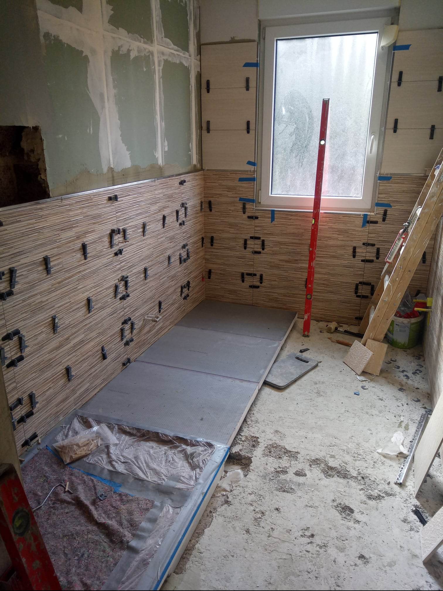 Łazienka w trakcie prac: częściowo ułożone płytki imitujące drewno na ścianach, poziomica oparta o okno, płyty podłogowe przygotowane do montażu, widoczne narzędzia i materiały budowlane.