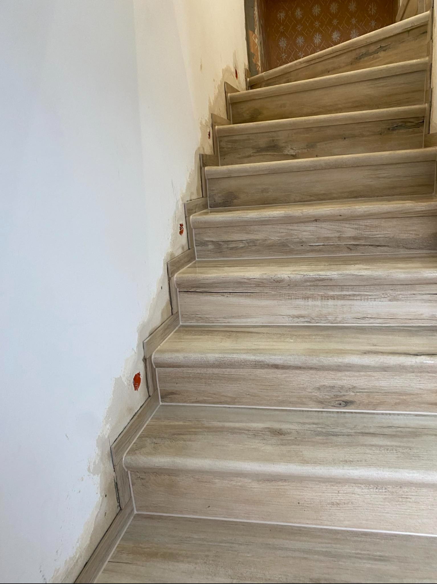 Schody wyłożone płytkami imitującymi drewno, widoczne ślady prac na ścianie, w tle fragment starej tapety.