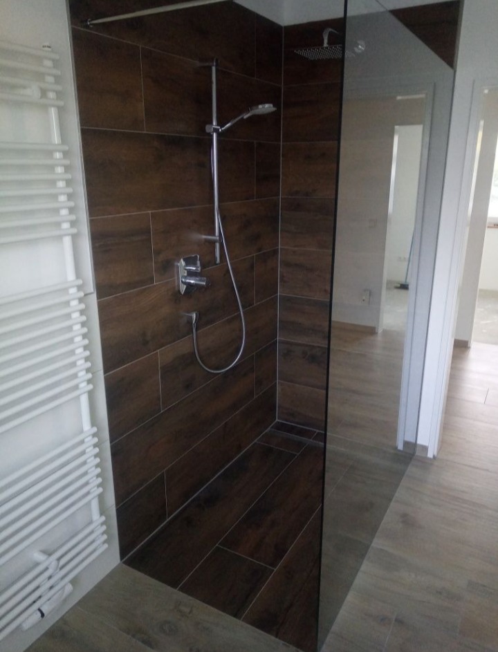 Nowoczesna kabina prysznicowa walk-in z ciemnobrązowymi płytkami imitującymi drewno, szklaną ścianą i grzejnikiem drabinkowym po lewej stronie.