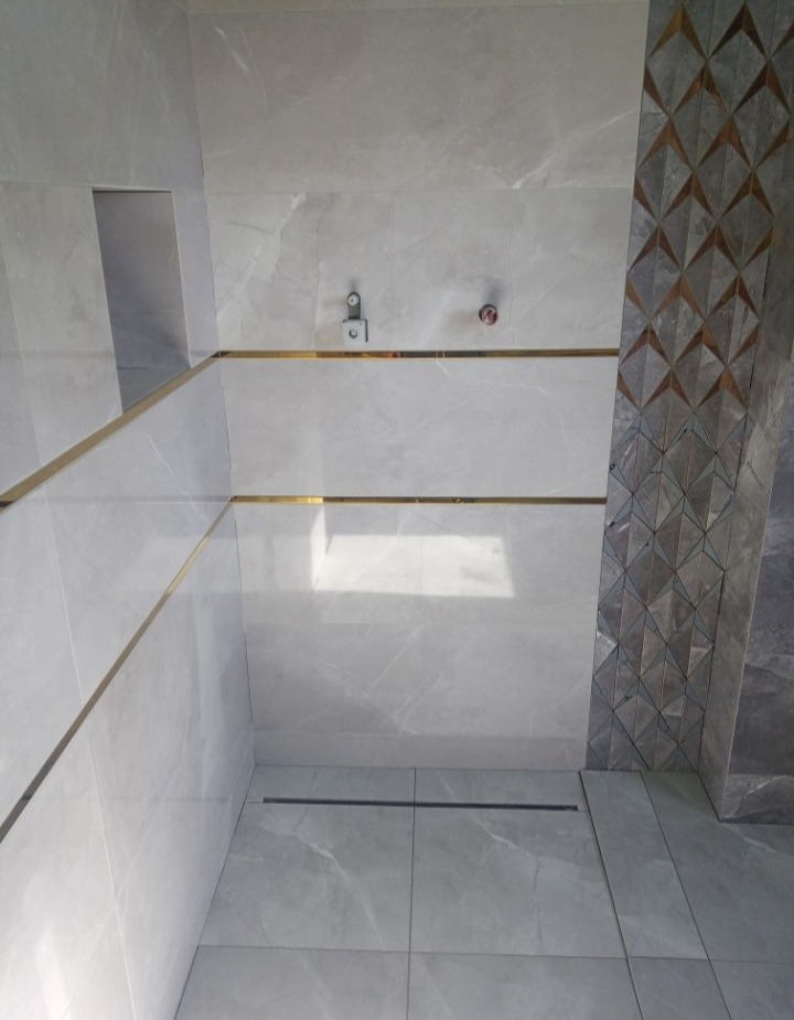 Wykończona łazienka z szarymi płytkami imitującymi marmur, złotymi listwami dekoracyjnymi i pionowym pasem płytek z geometrycznym wzorem. Widoczny odpływ liniowy w podłodze i wnęka w ścianie.