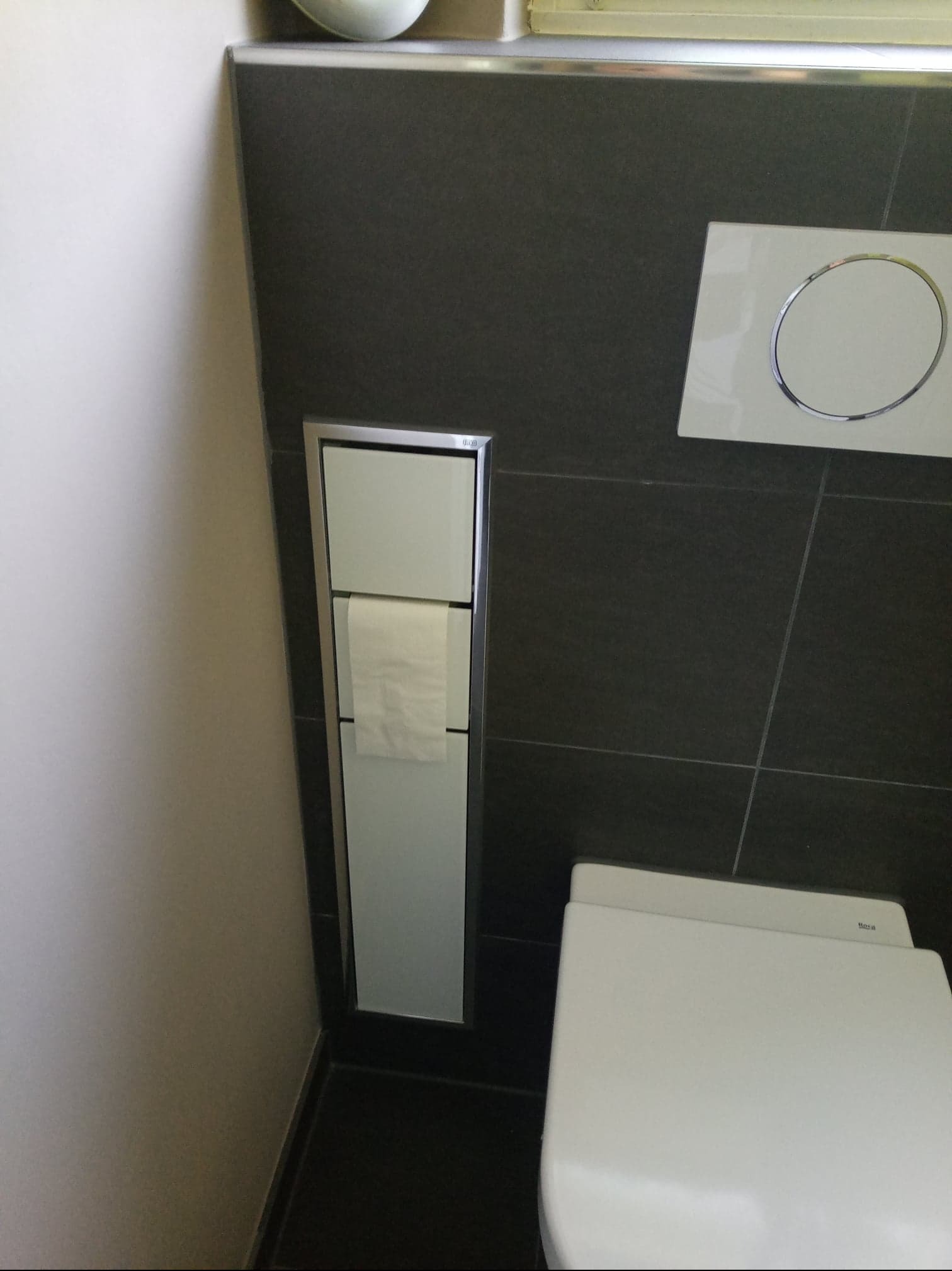 Nowoczesna toaleta podwieszana z czarną płytką i wbudowanym pojemnikiem na papier toaletowy, widok z bliska na detale wykończenia.